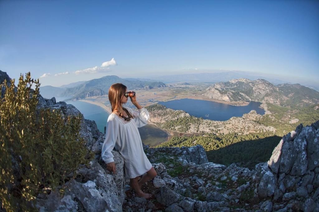 Une personne se tient sur un promontoire rocheux, d’où elle domine un paysage montagneux avec un lac.