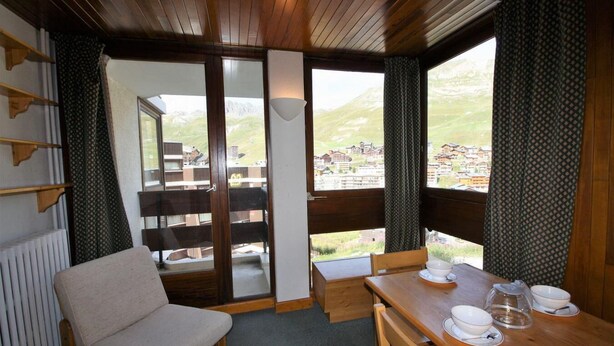 Una habitación acogedora con una mesa de madera lista para comer, una silla cómoda y vistas a un pueblo de montaña a través de amplios ventanales.