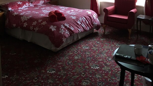 Un dormitorio con edredón estampado, una silla roja, una mesita con una taza y un control remoto, y una ventana con cortinas rojas.