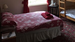 Un dormitorio con una cama individual, una mesita con una taza, un televisor montado en la pared y un ventanal con cortinas rojas.