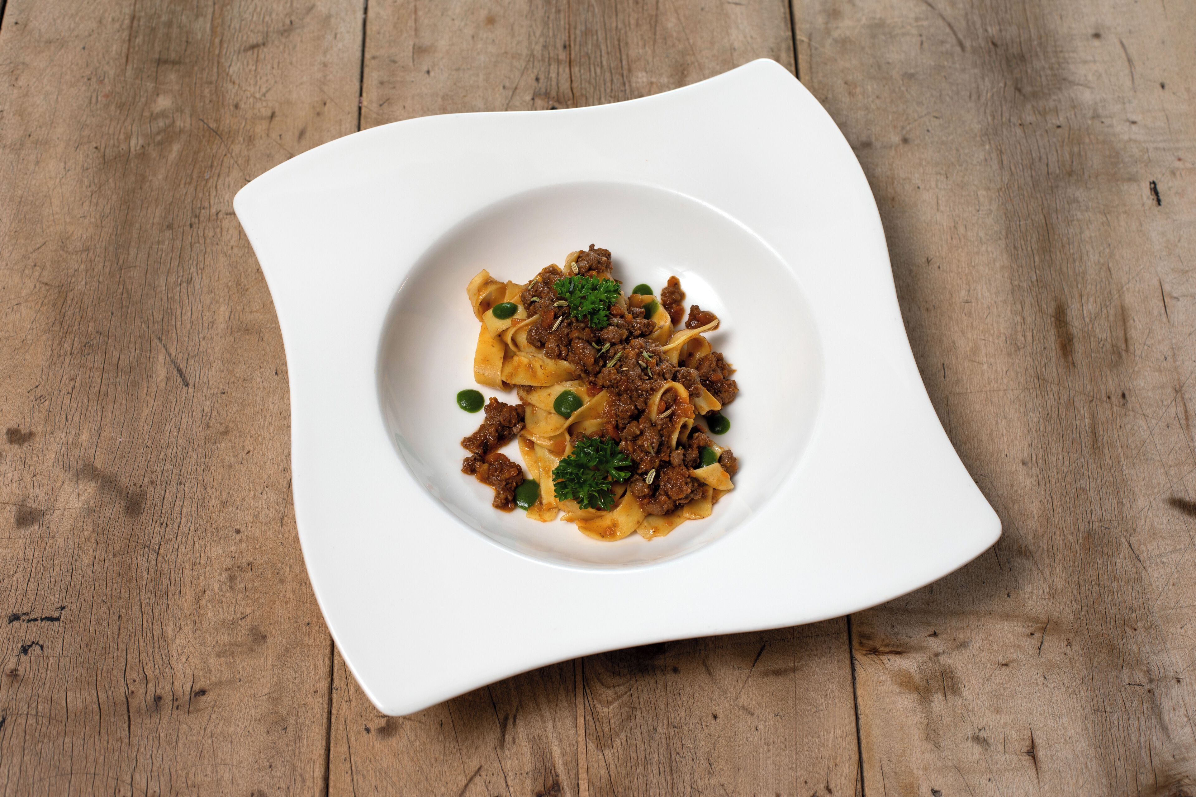 A square white plate with pasta and meat on a wooden table.