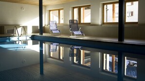 An indoor swimming pool with clear water, wooden ceiling, and windows with blinds. - Hotel Bodenhaus (Rheinwald)