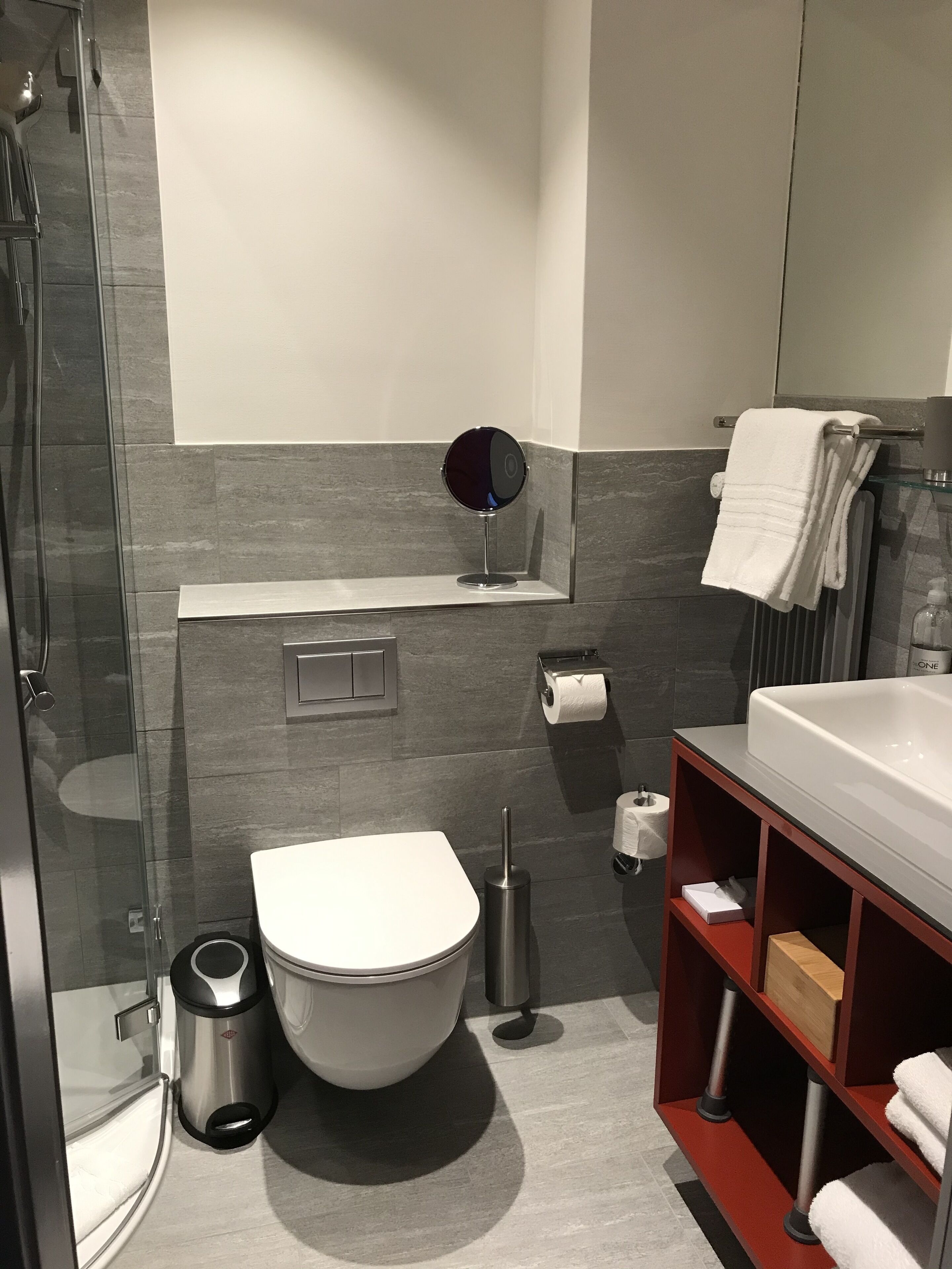 A modern bathroom with a glass shower enclosure, a white toilet, a round mirror, and a red vanity with a towel rack.