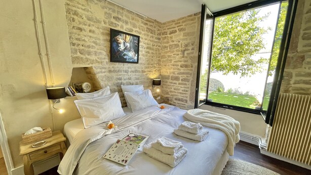 A bedroom with a stone wall, a large window, a bed with white linens, and a painting above the bed. - Mon Coeur House and Garden designed by Stay in Beaune is a unique property  (Beaune)