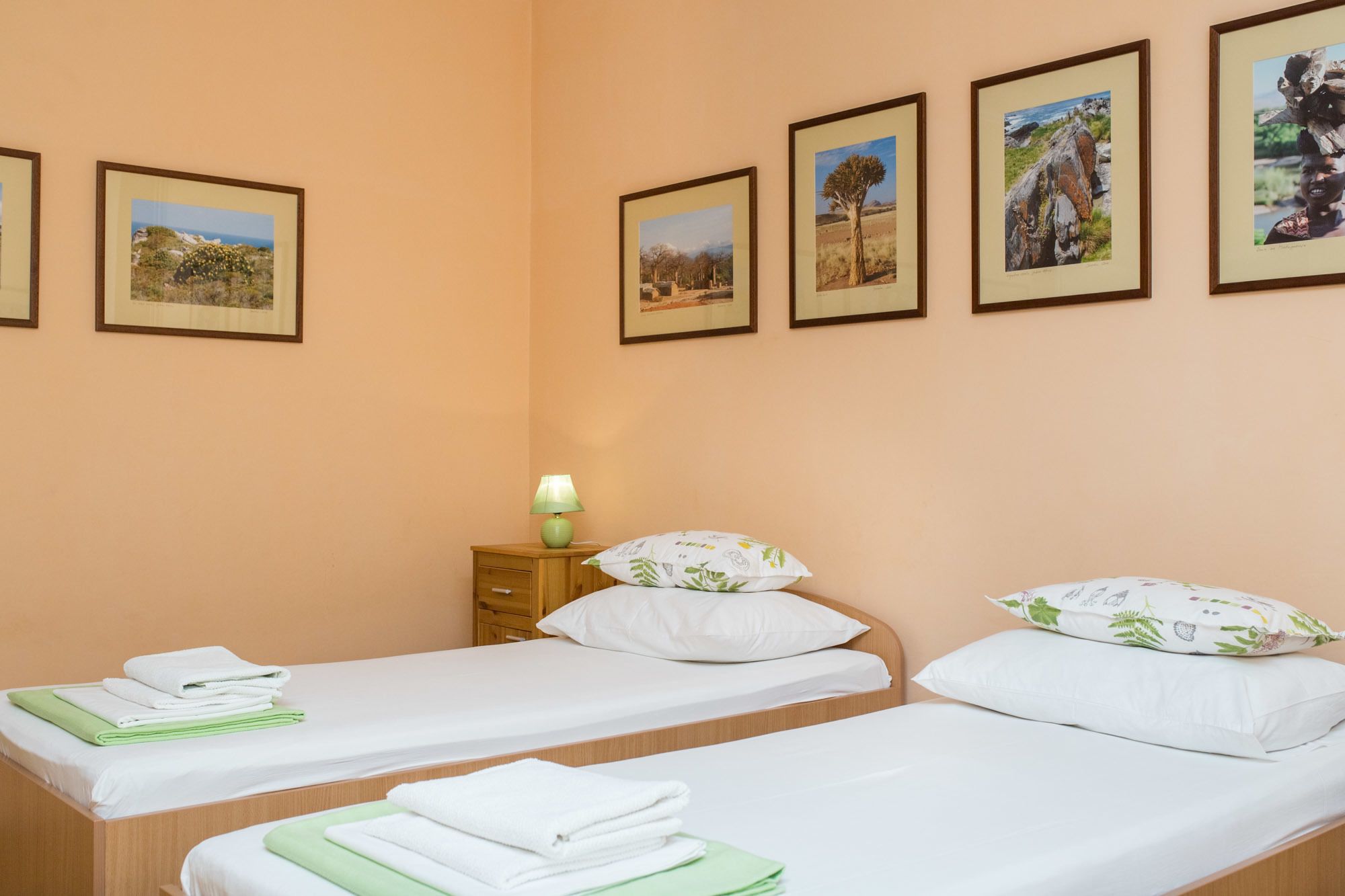 Une chambre d’hôtel avec deux lits simples, chacun agrémenté de draps blancs et de serviettes vertes. Des tableaux représentant des paysages, encadrés, ornent le mur.