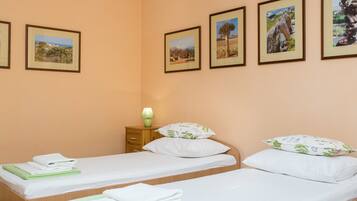 Une chambre d’hôtel avec deux lits simples, chacun agrémenté de draps blancs et de serviettes vertes. Des tableaux représentant des paysages, encadrés, ornent le mur.