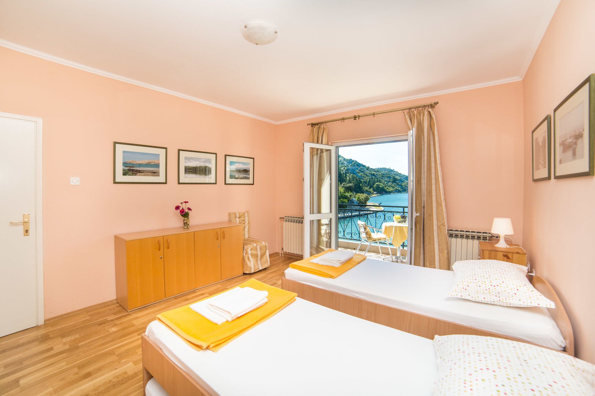 A hotel room with two beds, a wooden dresser, a view of the sea, and framed pictures on the wall.