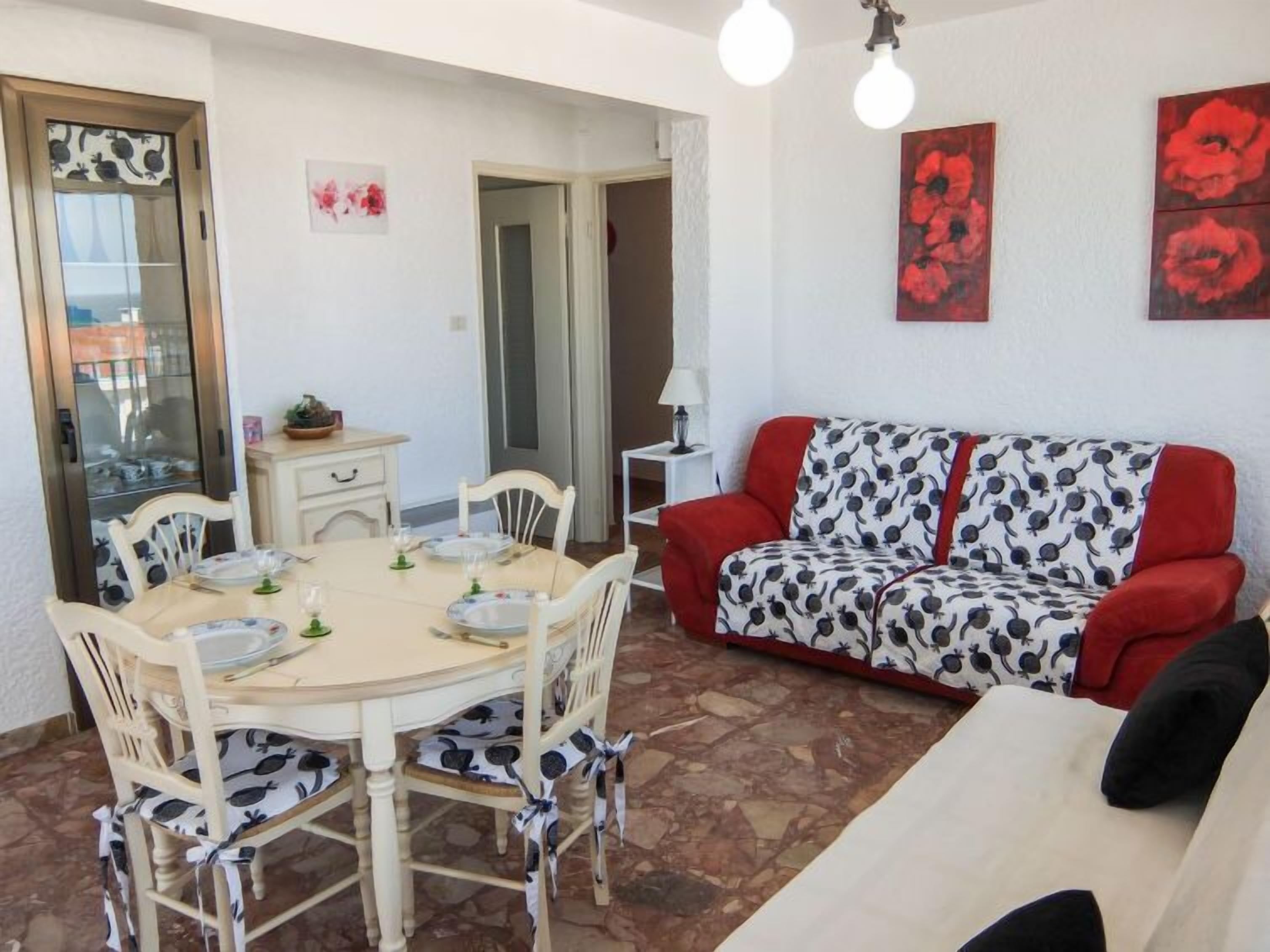 A dining area with a round table and chairs, a red and white sofa, and a small side table with a lamp.