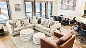 A modern living room with a sectional sofa, ottomans, a leather armchair, a dining area with a table and chairs, and large windows with blinds.