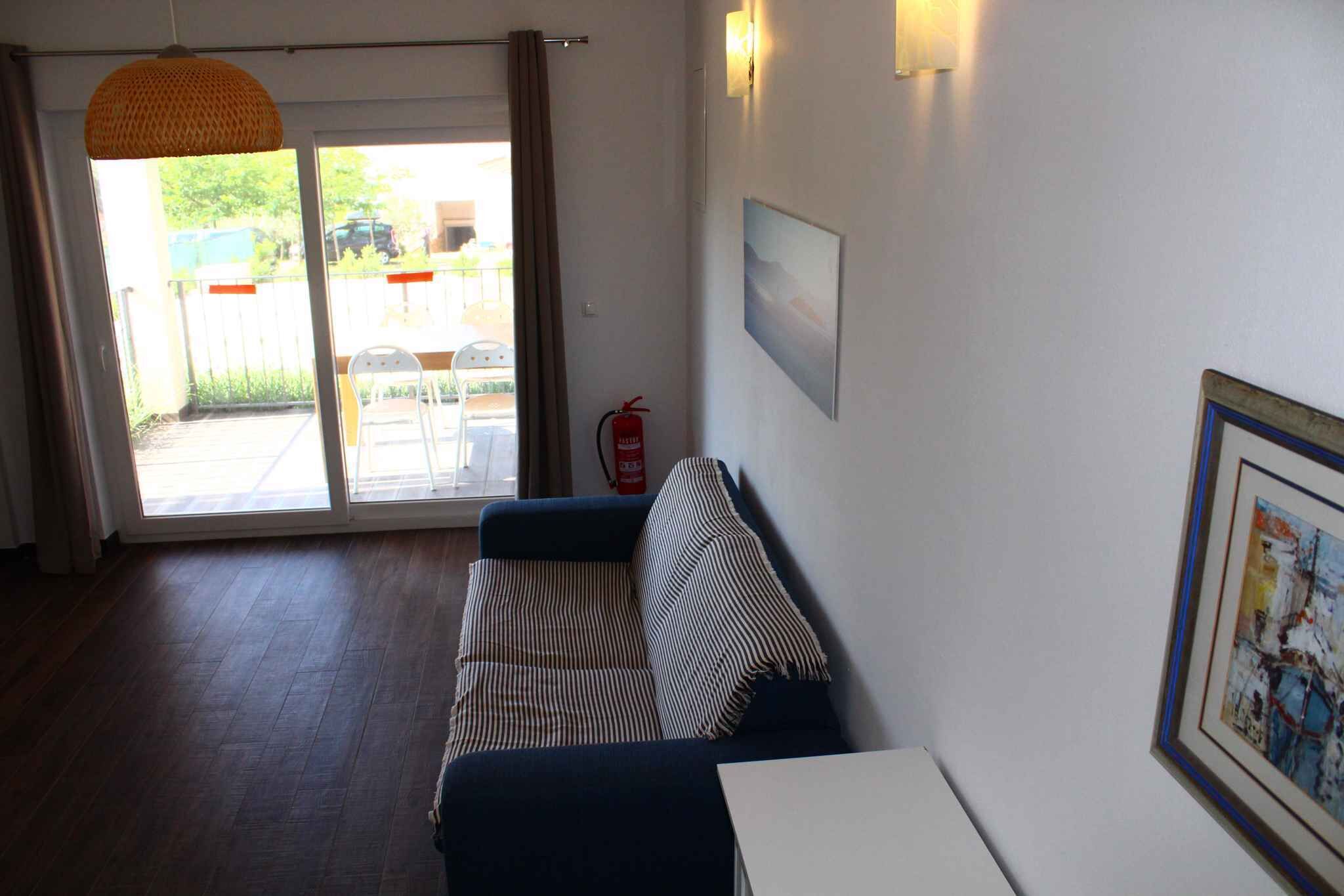 A room with a sofa, a table, a fire extinguisher, and a door leading to an outdoor area with chairs and a fence.