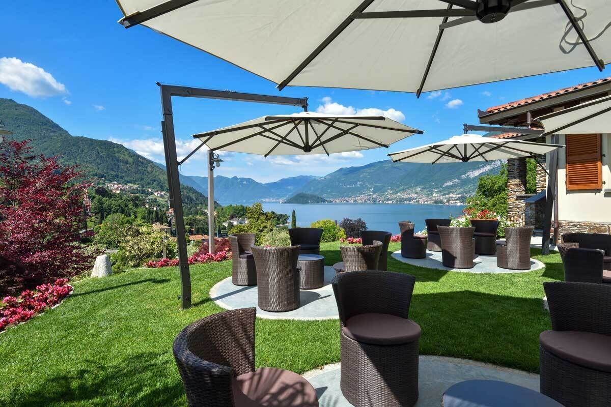 An outdoor seating area with wicker furniture and umbrellas, overlooking a scenic mountainous landscape and a body of water.