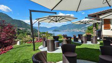 An outdoor seating area with wicker furniture and umbrellas, overlooking a scenic mountainous landscape and a body of water.