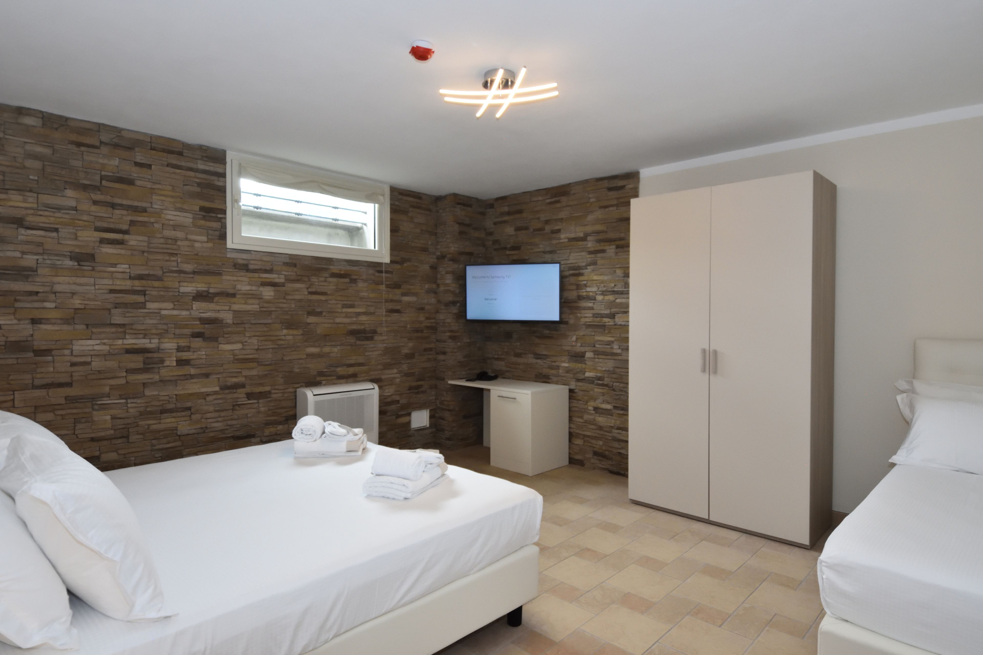 A modern hotel room with a stone wall, a flat-screen TV, a wardrobe, and a bed with white linens.