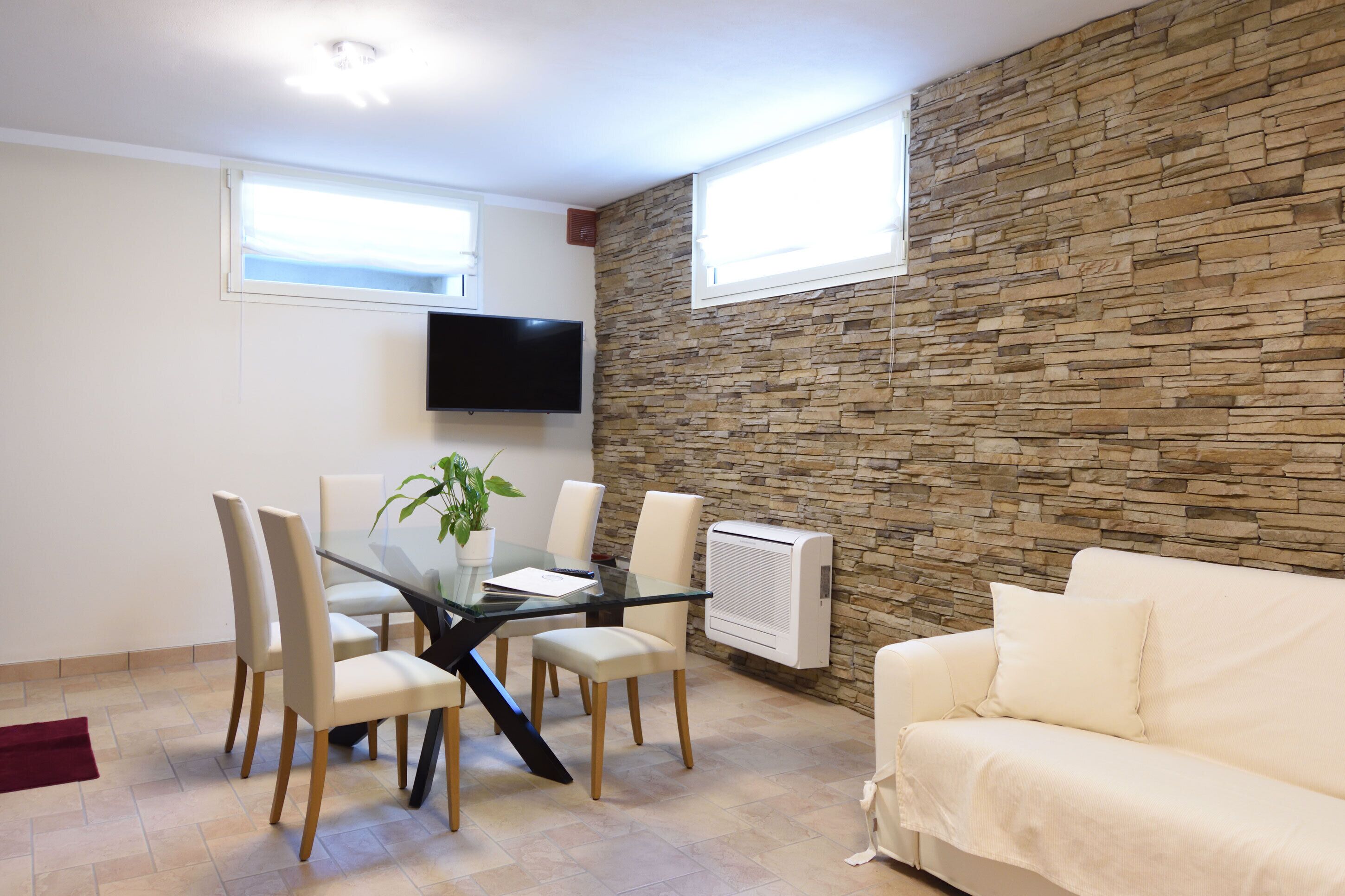 A modern living room with a stone wall, a glass dining table with chairs, a white sofa, and a mounted TV.