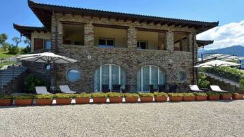 A two-story stone building with a balcony, outdoor seating with umbrellas, and a gravel driveway.
