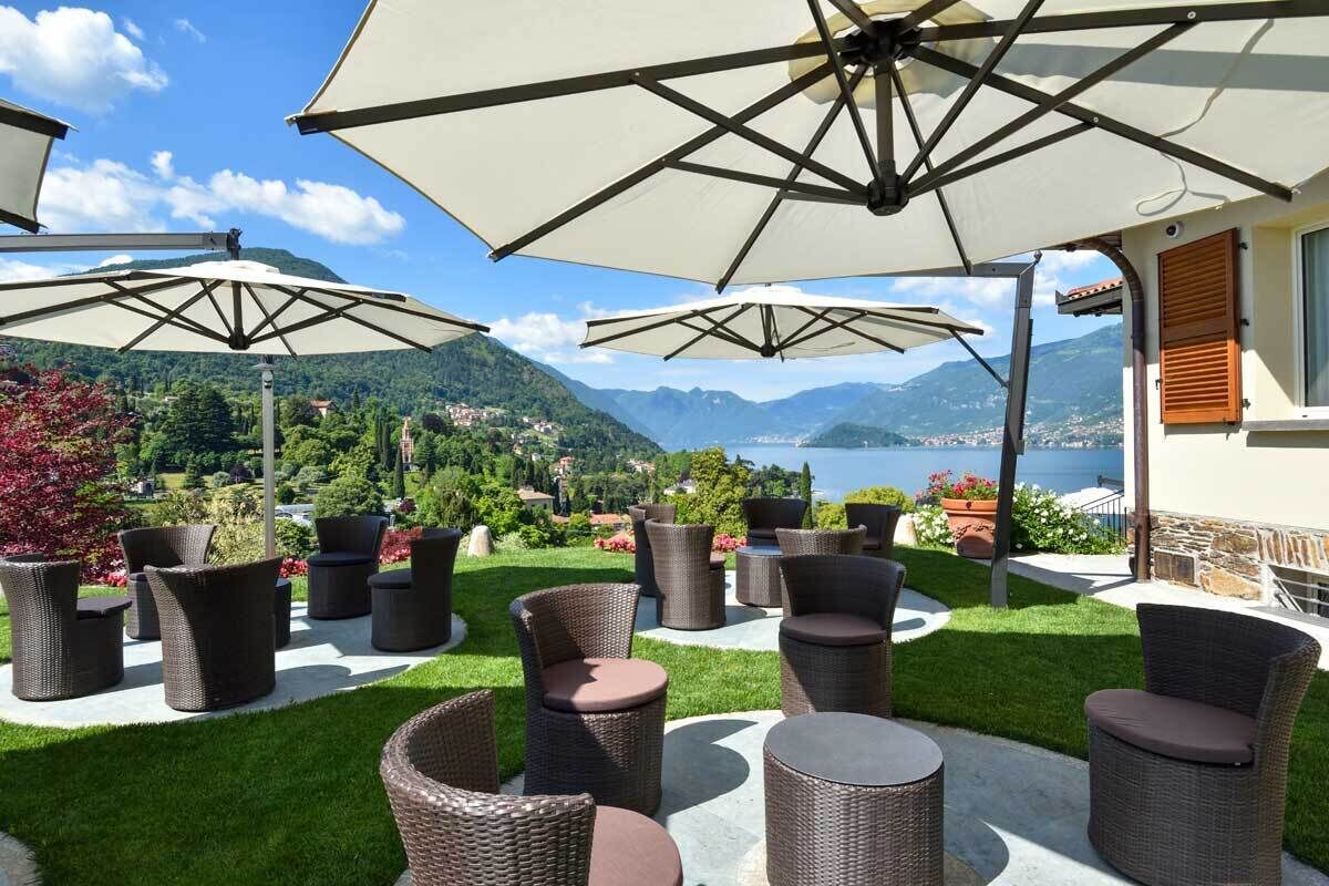 An outdoor seating area with wicker furniture and large umbrellas, overlooking a mountainous landscape and a body of water.