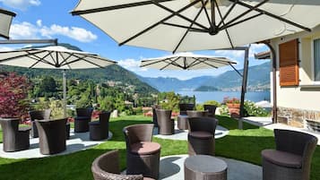 An outdoor seating area with wicker furniture and large umbrellas, overlooking a mountainous landscape and a body of water.