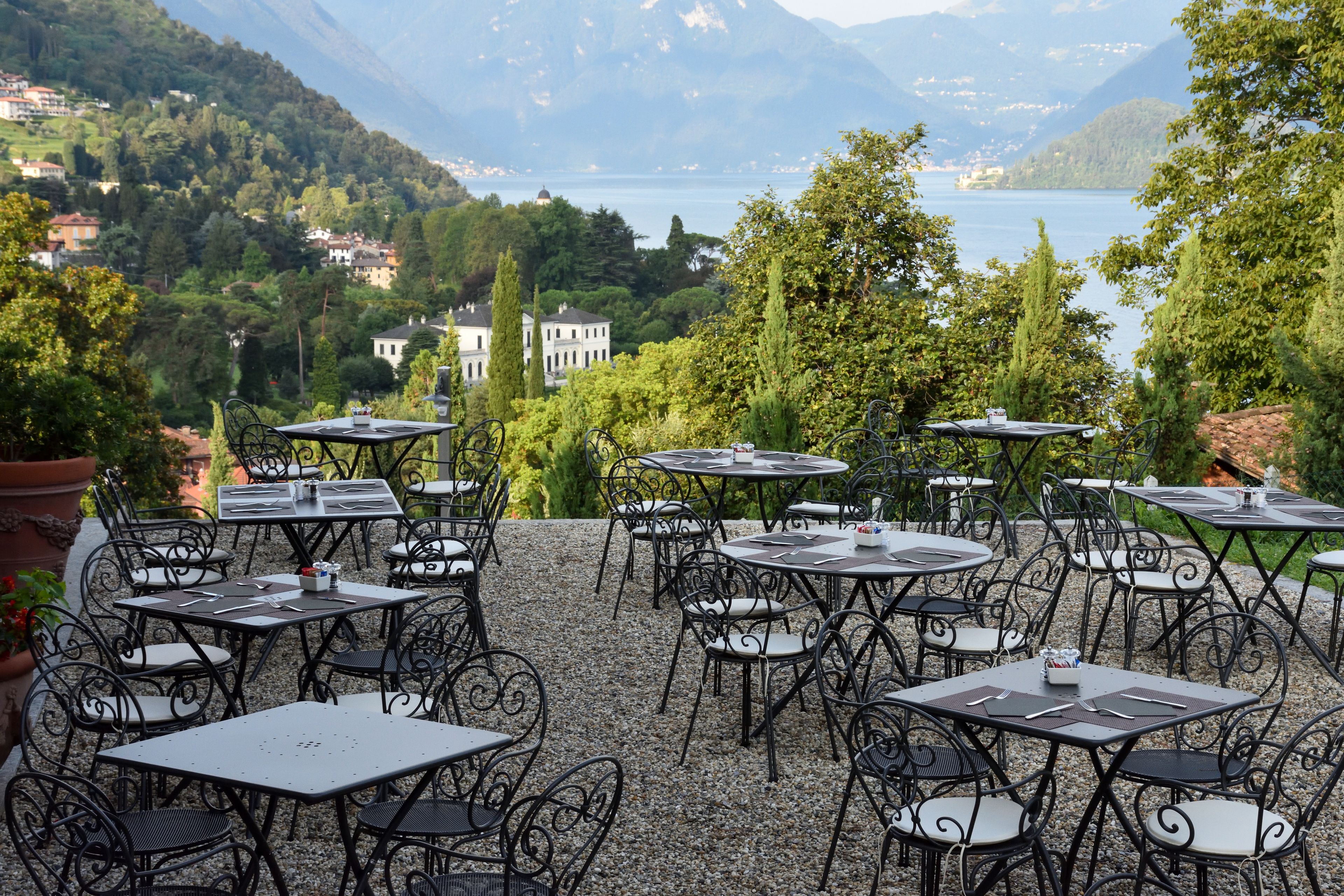 Tische und Stühle im Außenbereich auf einer Terrasse mit Blick auf einen See und Berge im Hintergrund.