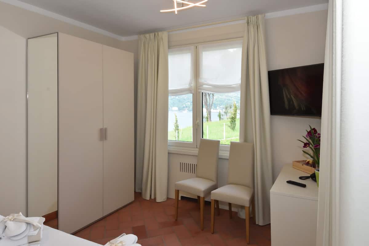 Hotellihuone, jossa on suuri vaatekaappi, kaksi tuolia, pieni pöytä, jossa on maljakko täynnä kukkia, ja ikkuna, josta avautuu näkymä vehreään maisemaan ja järvelle.