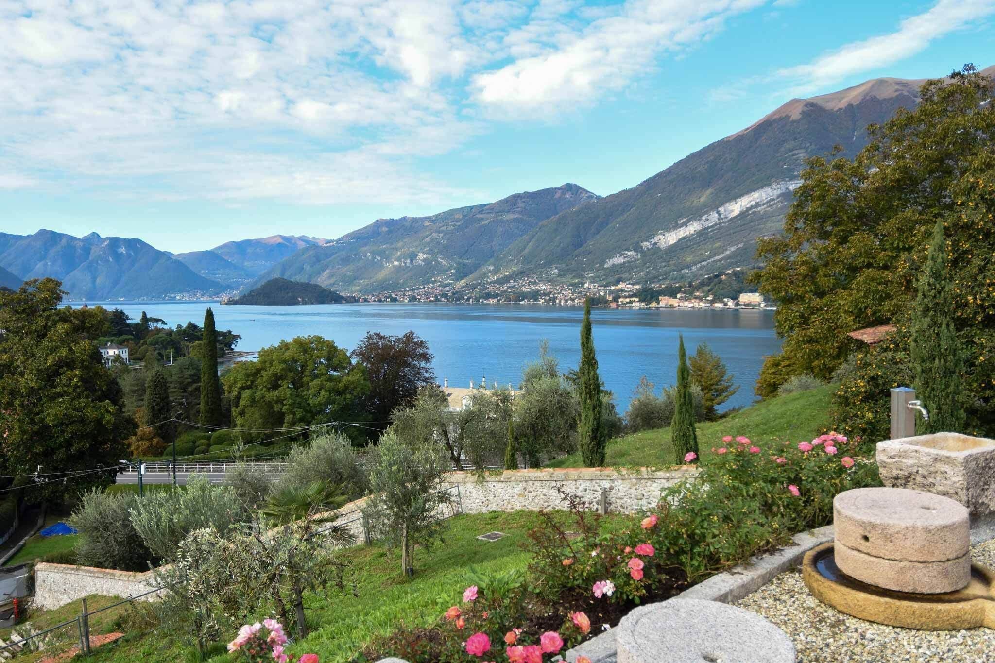 Una vista panorámica de un lago con montañas al fondo, un pozo de piedra y un muro de piedra adornado con flores.