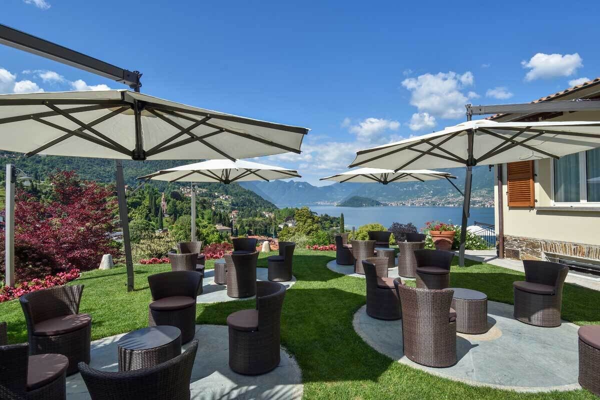 Un patio con mobiliario de mimbre y sombrillas grandes, con vistas a un paisaje montañoso y un lago.