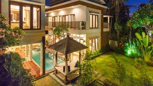 Ein modernes, zweistöckiges Haus mit Swimmingpool, überdachter Terrasse und üppiger tropischer Bepflanzung.