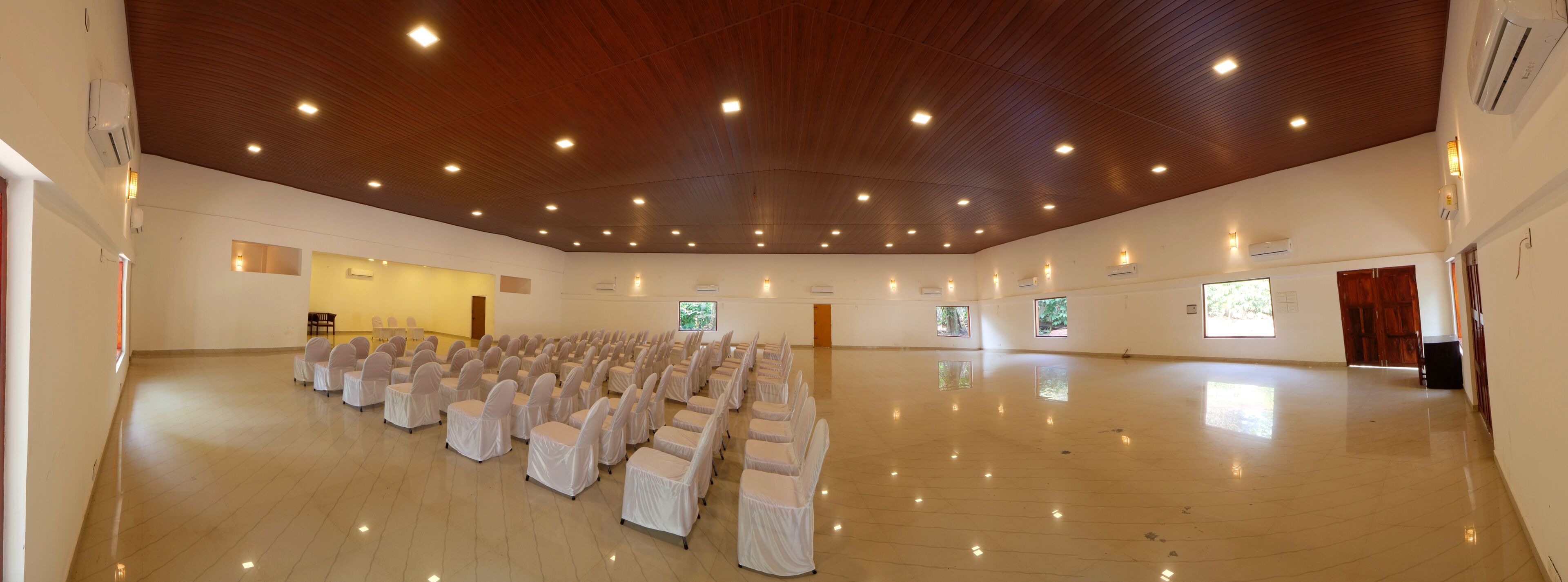 A spacious hall with rows of chairs covered in white cloth, wooden ceiling with recessed lighting, and large windows allowing natural light.
