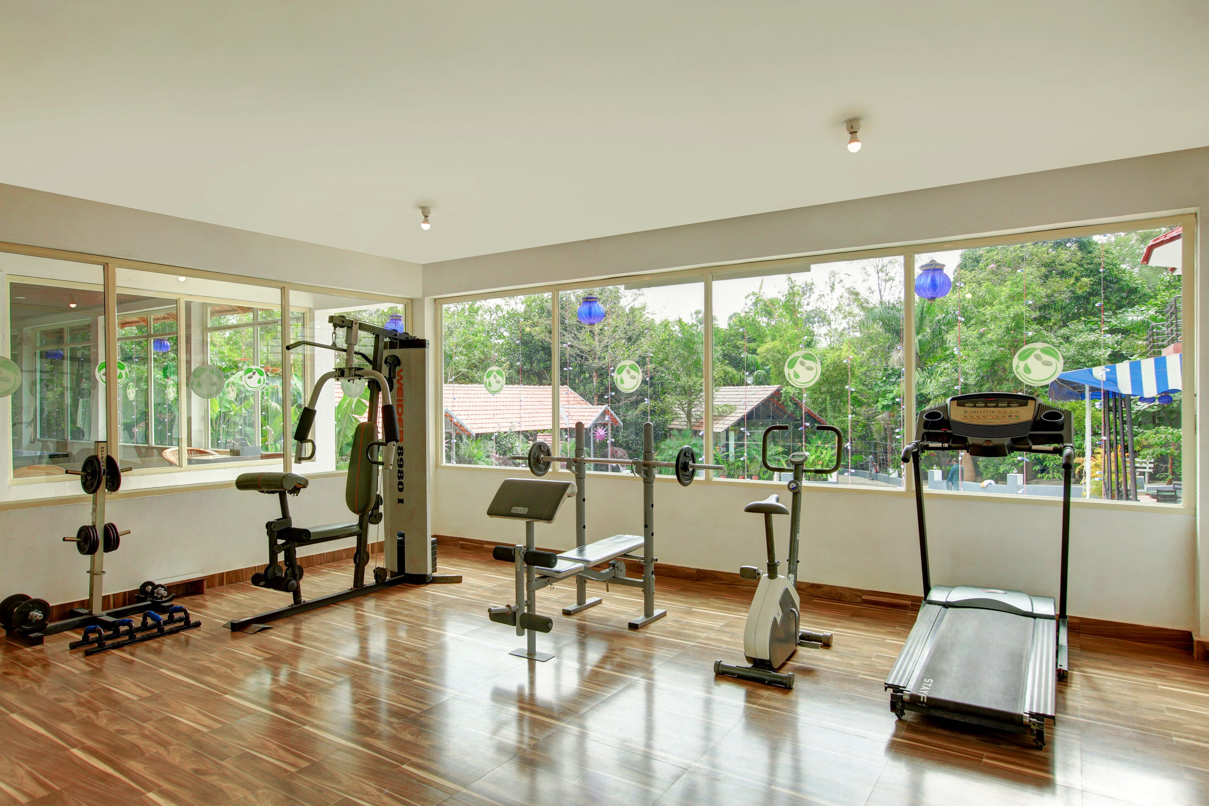 A gym with various exercise equipment, large windows, and a view of trees and buildings outside.