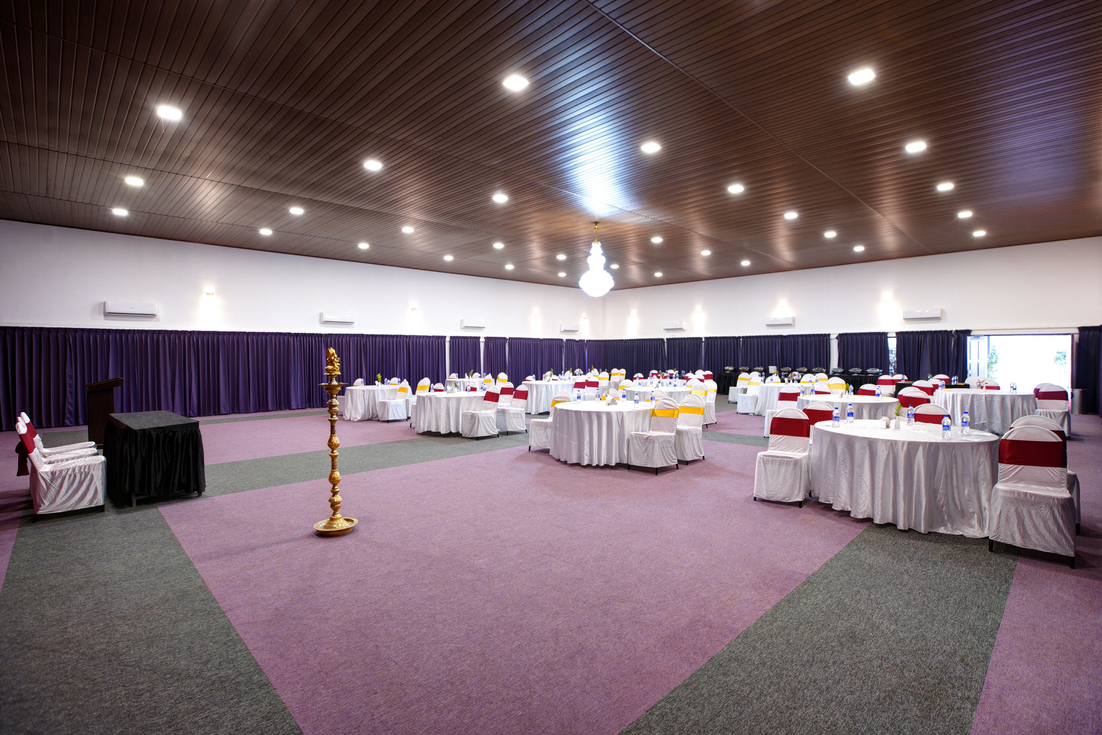 A large hall with round tables and chairs, a podium, and a decorative golden pole.