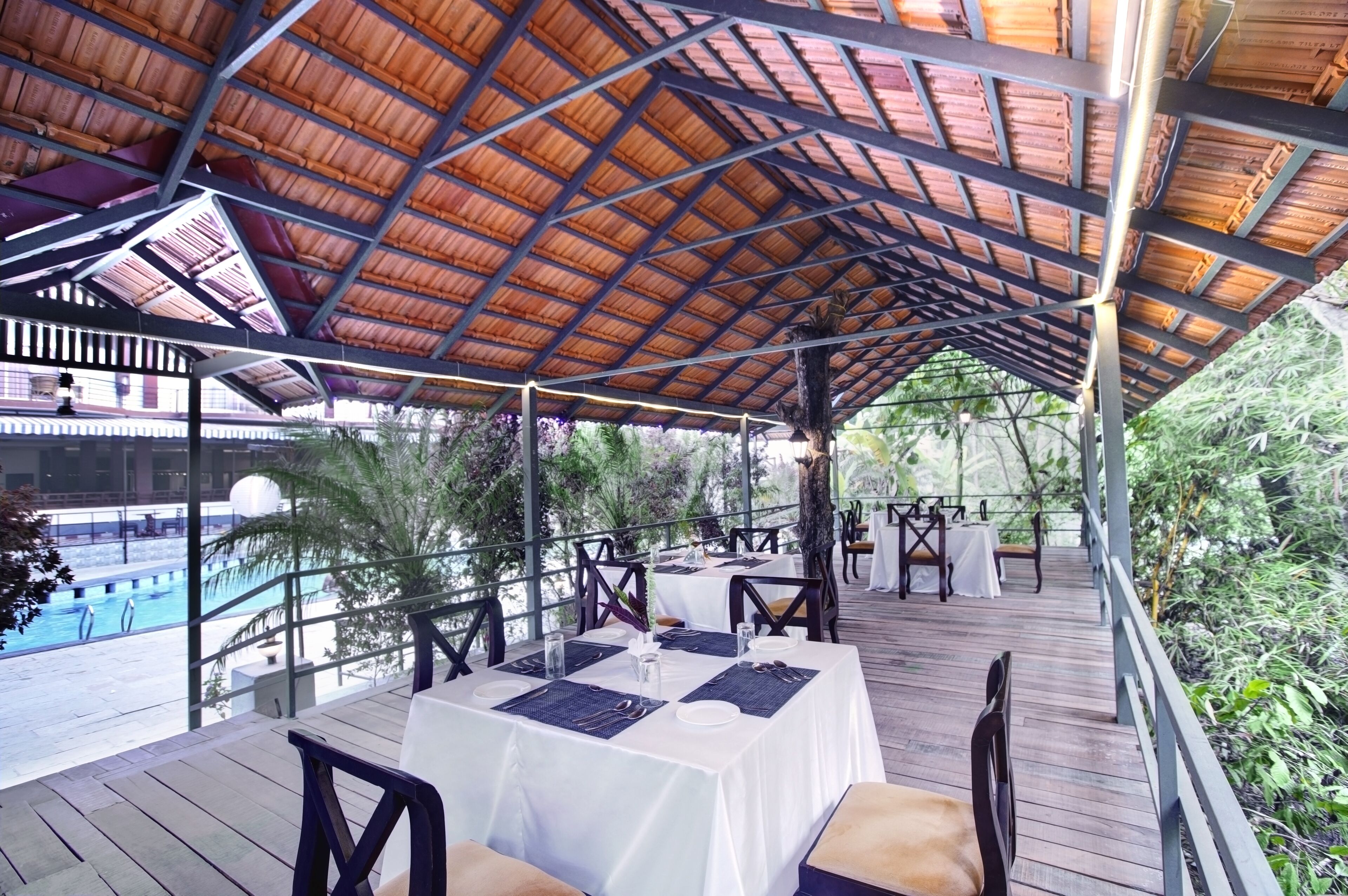An outdoor dining area with tables set for two, under a wooden pergola with a view of a pool.
