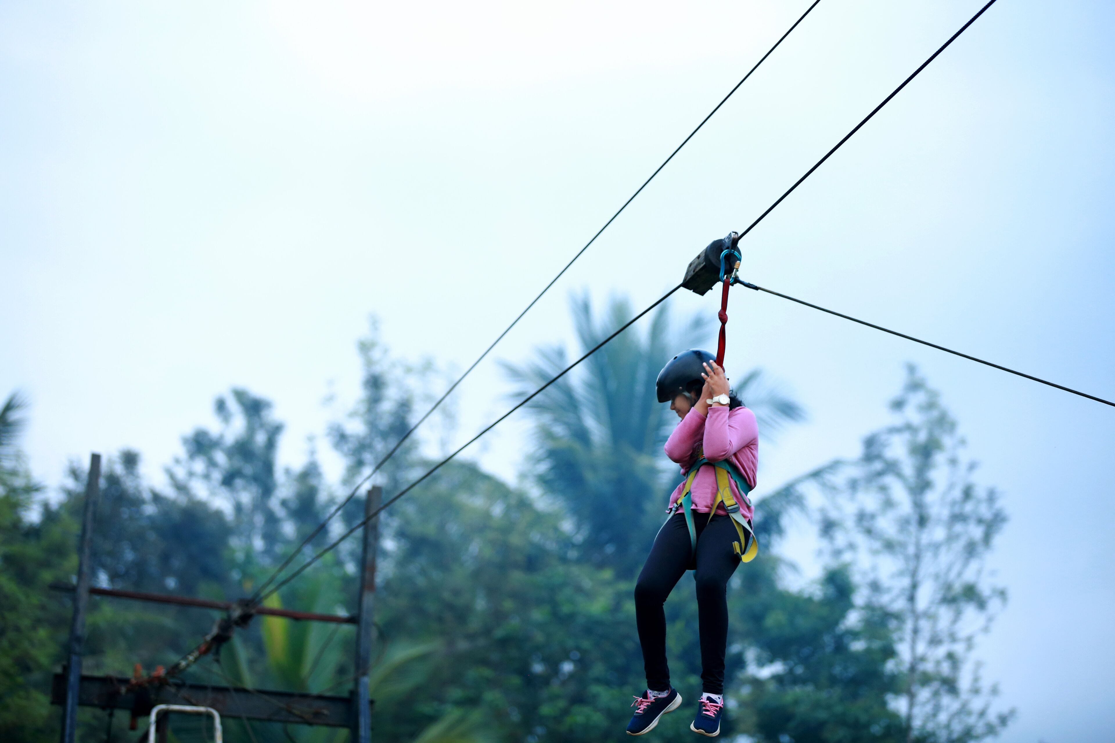 A person zip-lining with safety harnesses and helmet, surrounded by trees and a clear sky.