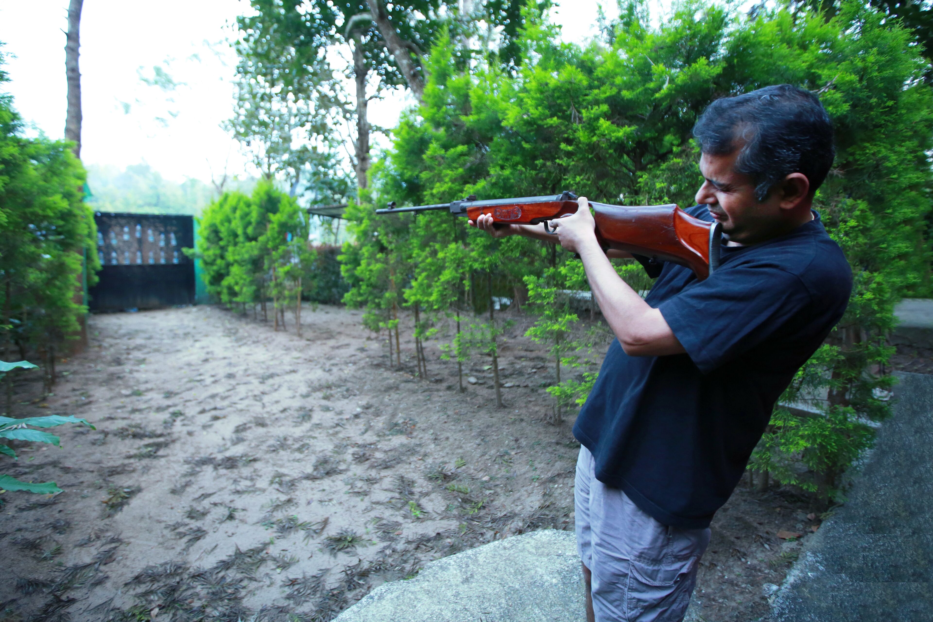 A person aiming a rifle in an outdoor setting with trees and a dirt path.