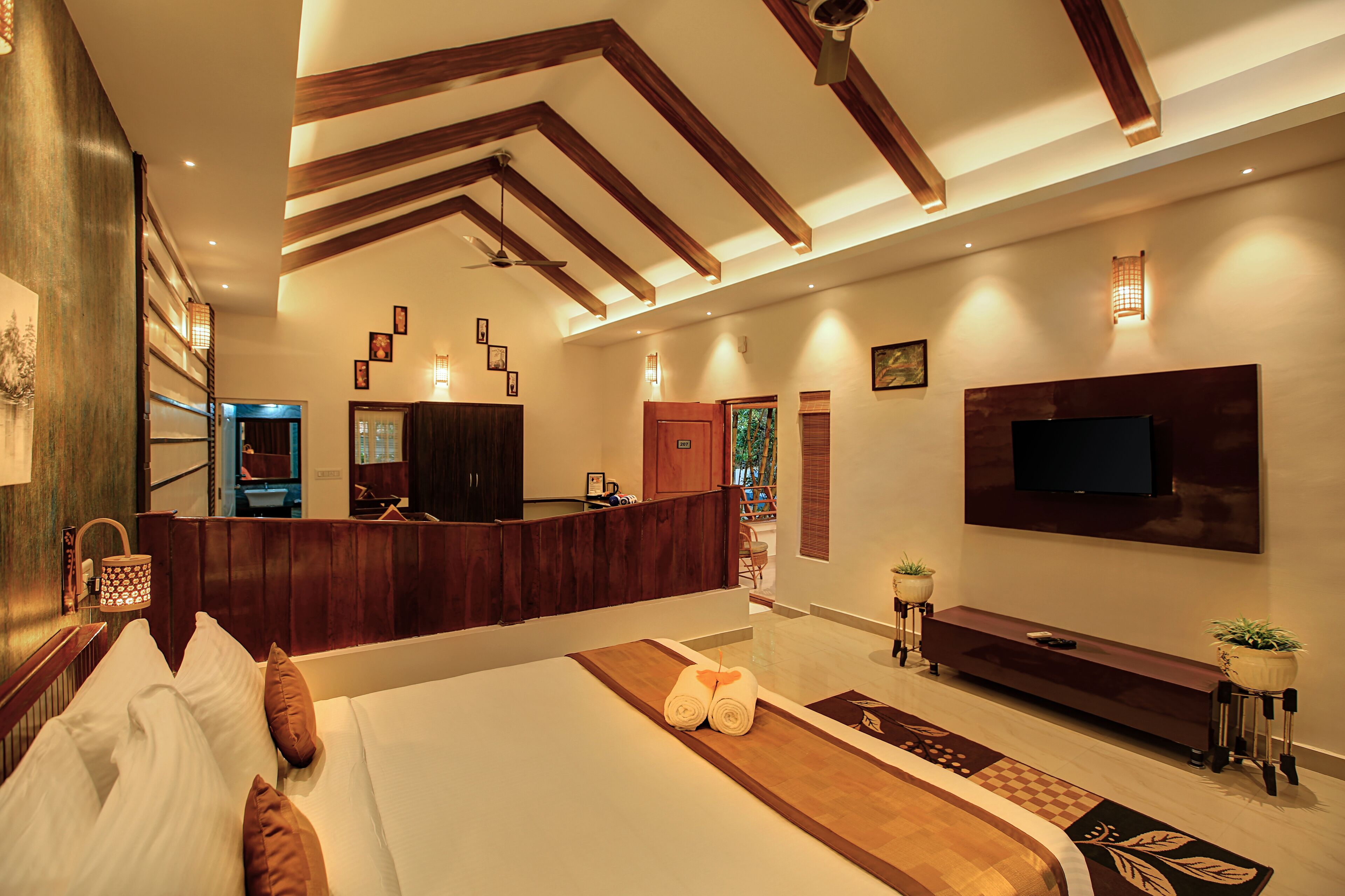 A modern hotel room with a large bed, a wooden reception desk, a flat-screen TV, and a ceiling fan.