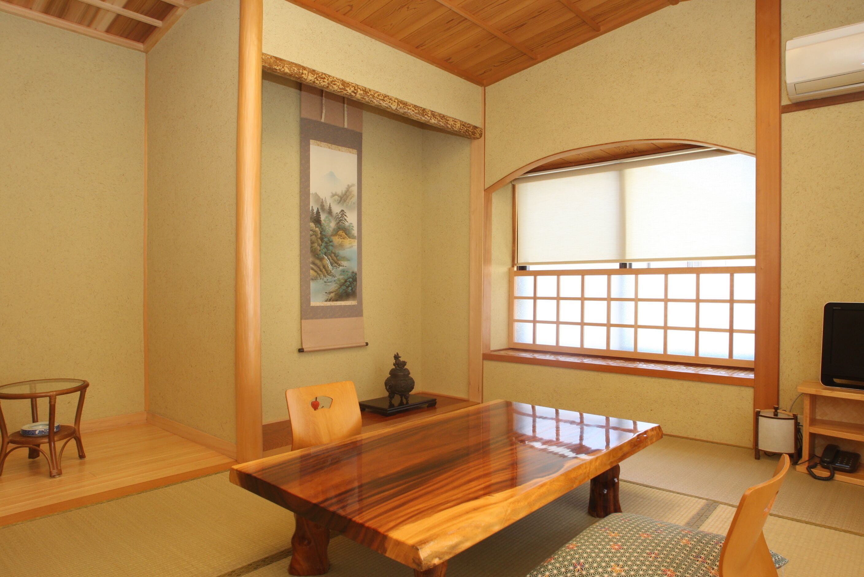 Una habitación tradicional japonesa con suelo de tatami, una mesa de madera para comer, una mesa baja con un cuenco, una ventana con celosía de papel shoji y una obra de arte colgada.