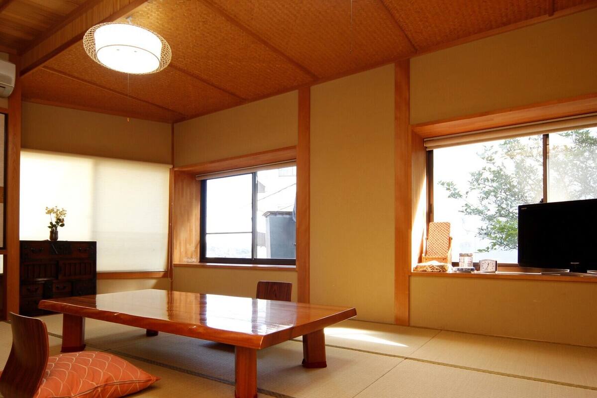 A traditional Japanese room with tatami flooring, a wooden table, a television, and a window with a view of greenery.