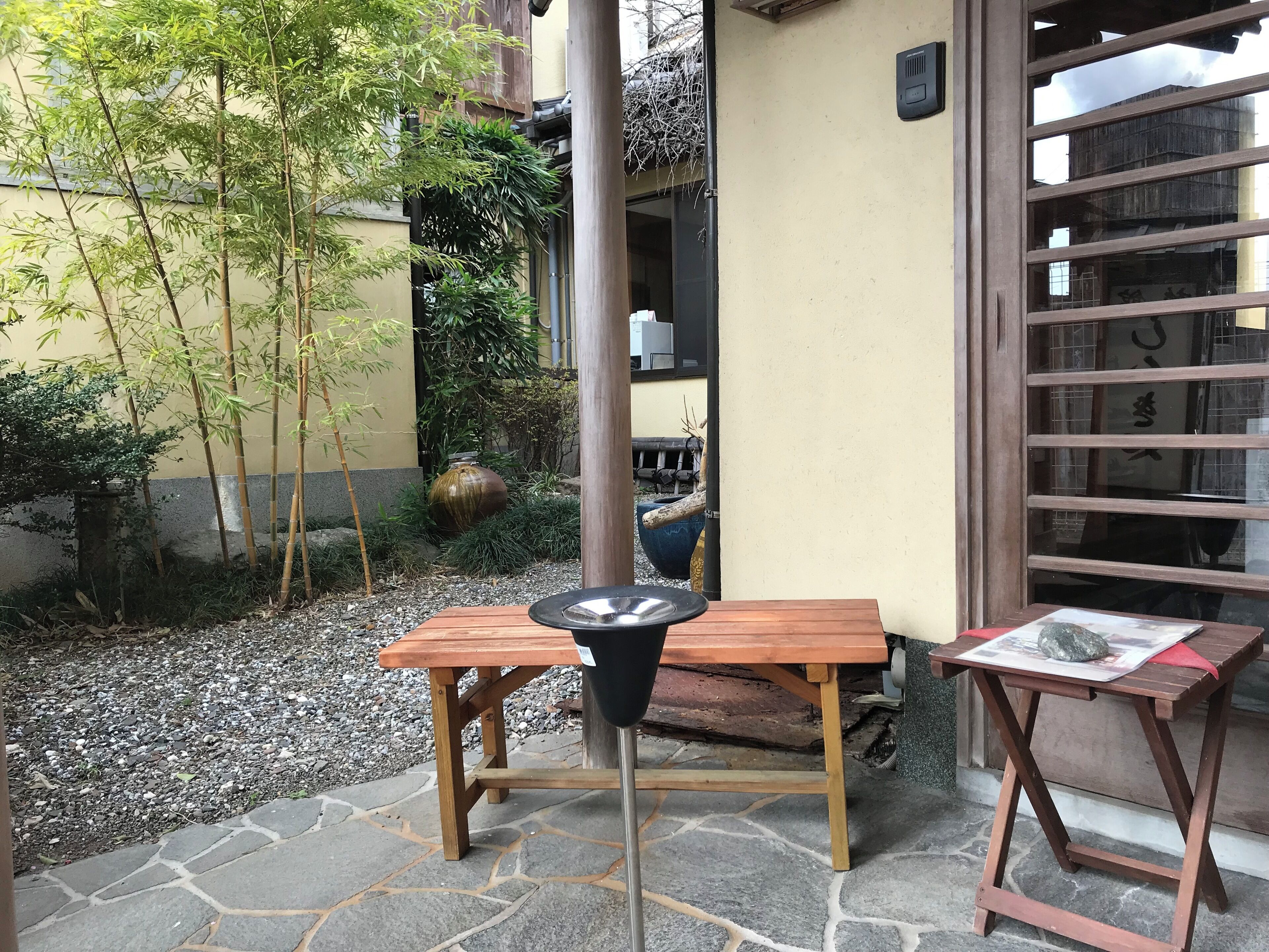 Una mesa de madera con un objeto negro encima, un patio con baldosas de piedra, plantas de bambú y un edificio con una verja de madera.