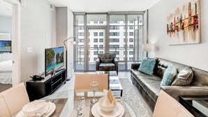 A modern living room with a sofa, a dining table set for two, a television, and a large window offering a view of a building. - Pet-friendly 1BR Den w/ Pool & Spa | nr Glen Echo Park (Bethesda)