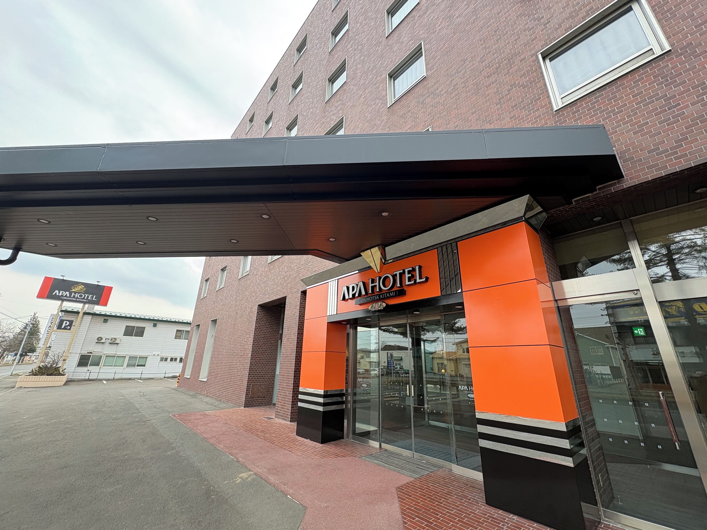 Entrada de hotel con diseño moderno, destacando una fachada prominente en naranja y negro, puertas de cristal amplias y un corredor cubierto.