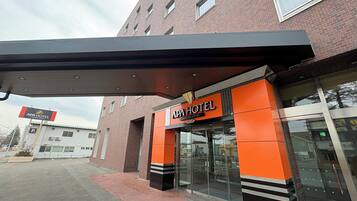 Entrada de hotel con diseño moderno, destacando una fachada prominente en naranja y negro, puertas de cristal amplias y un corredor cubierto.