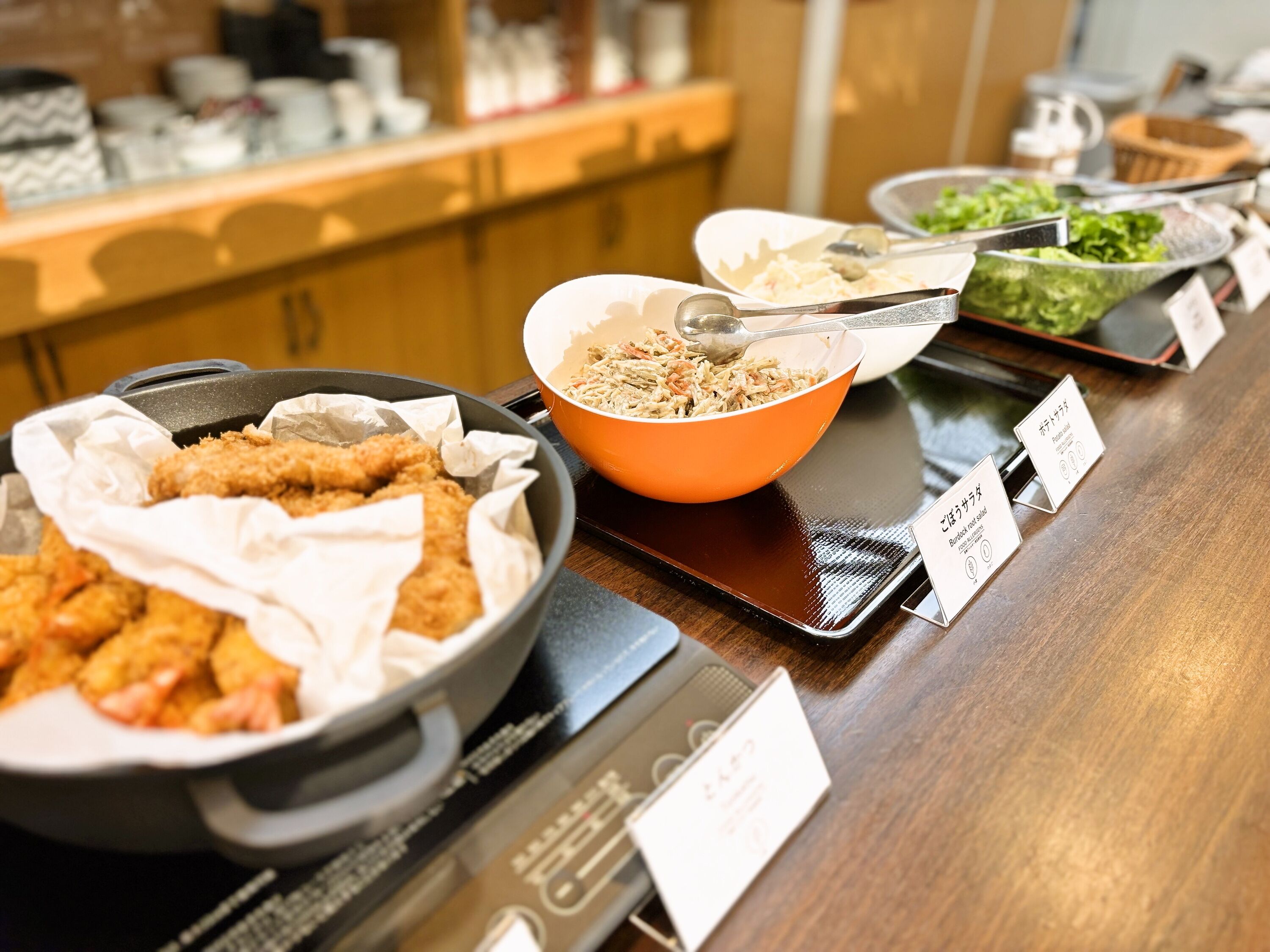 Un buffet proposant divers plats, notamment des plats frits et des salades, disposés sur une table en bois et accompagnés d’étiquettes.