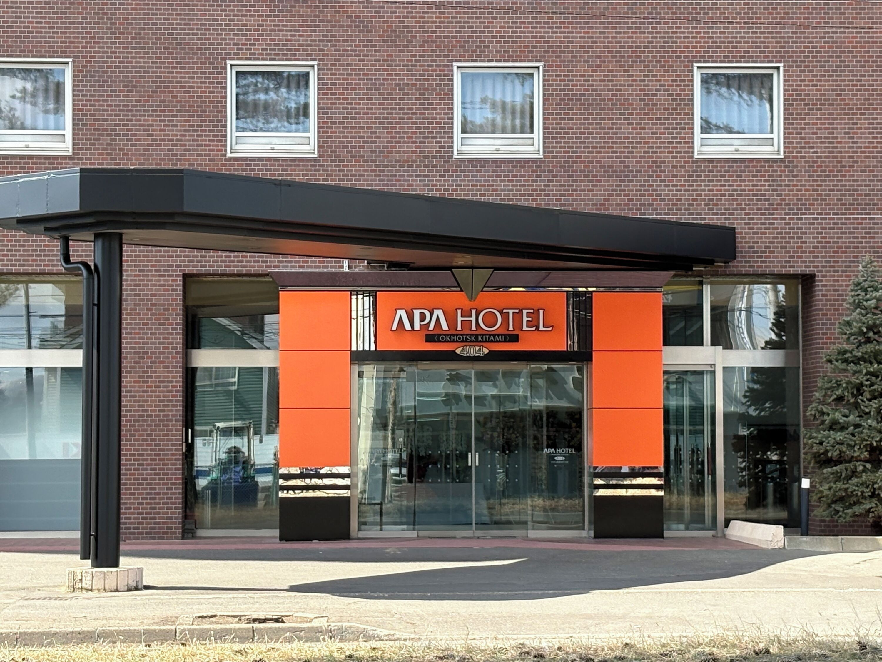 A brick building with a glass entrance, an orange sign reading 'APA HOTEL,' and a covered entrance area.