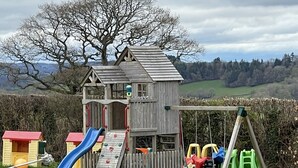 Ein Spielhäuschen aus Holz mit Rutsche und Kletterwand, umgeben von einer Schaukel und einem Picknicktisch.