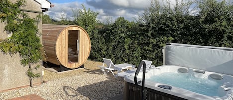 A hot tub and a sauna in a backyard with a brick patio, surrounded by greenery and a clear sky.