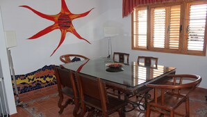 Een eetruimte met een glazen tafel, houten stoelen, een mozaïekwand en een raam met zonwering.