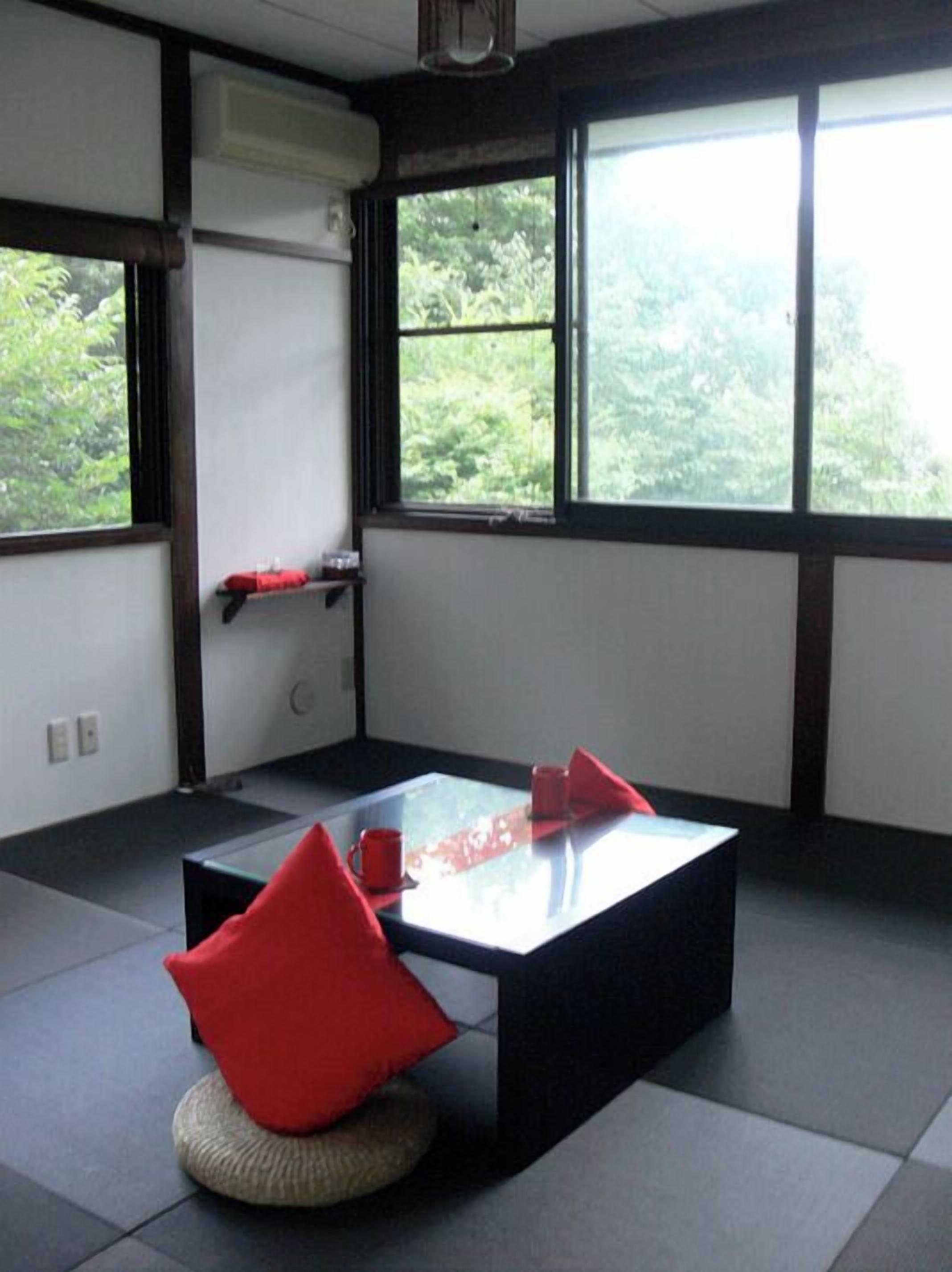 A room with tatami flooring, a low table with a cushion, and a view of greenery through large windows.