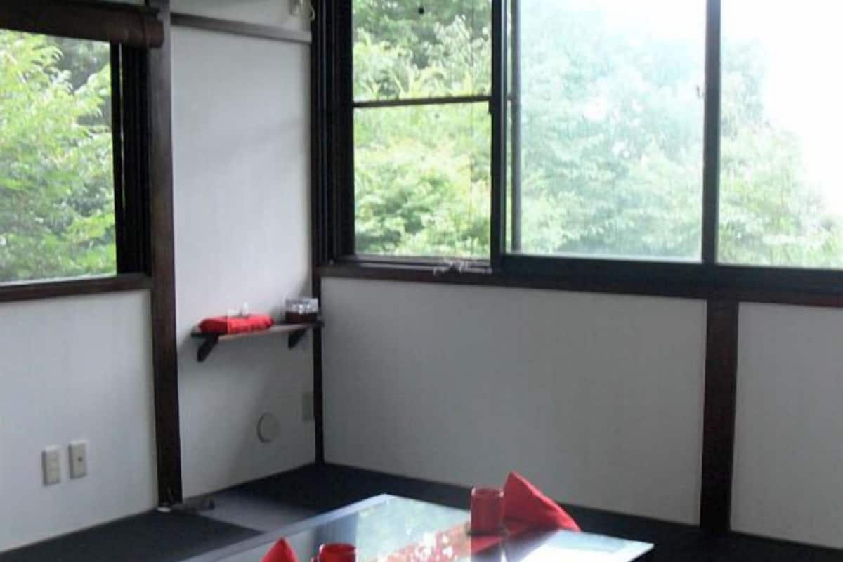 A room with tatami flooring, a low table with a cushion, and a view of greenery through large windows.