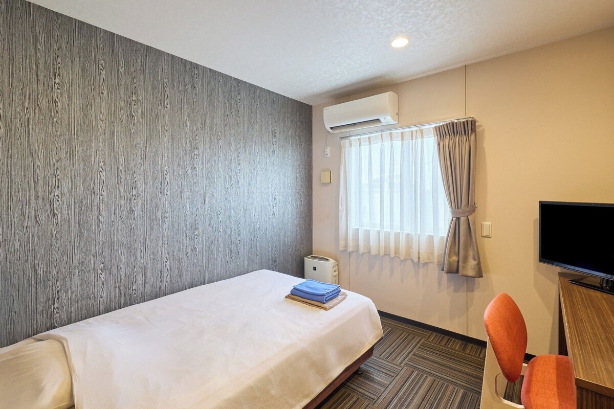 A hotel room with a bed, a TV, an air conditioning unit, a window with curtains, and a carpeted floor.