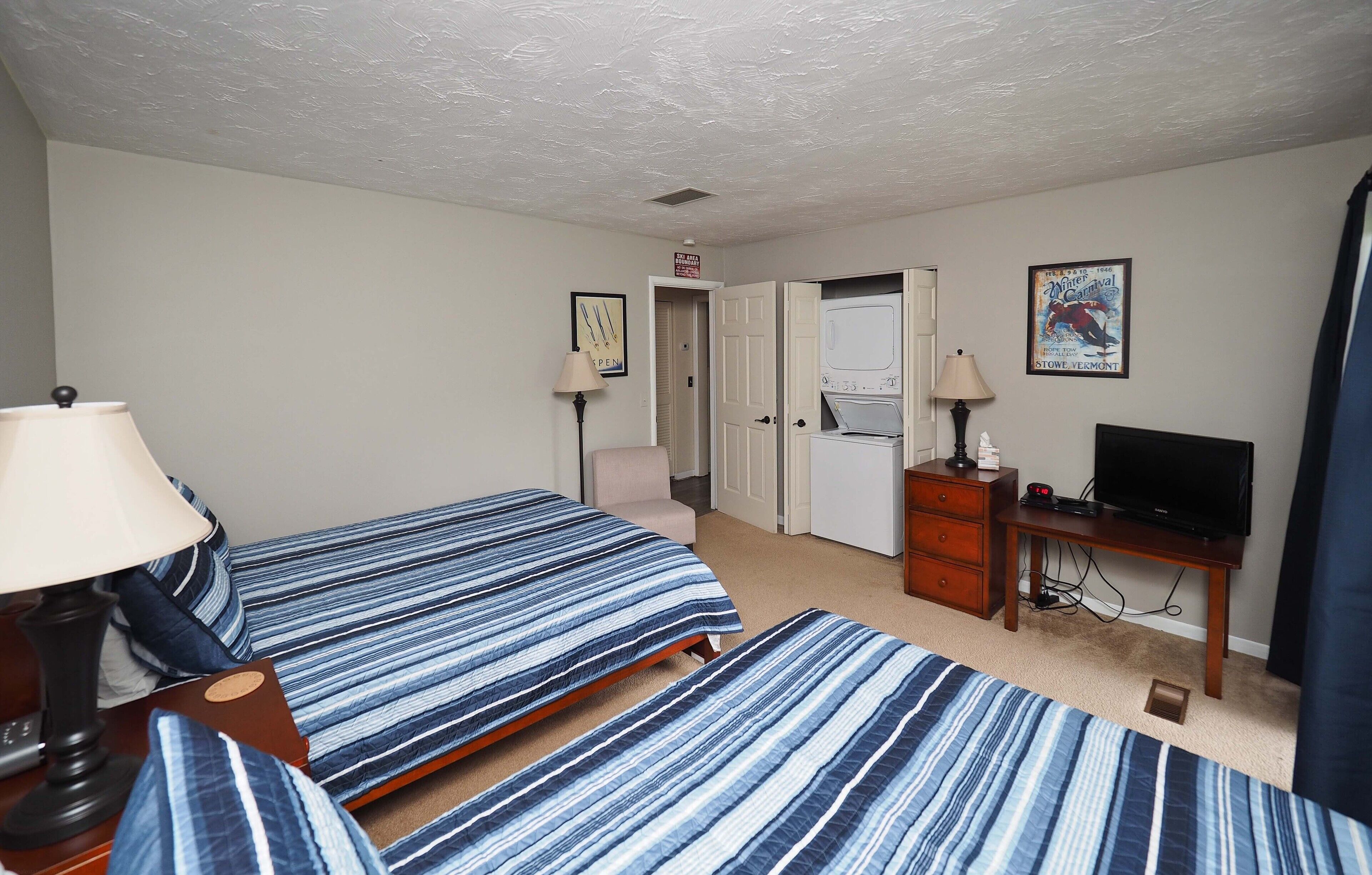 A hotel room with two beds, a desk with a television, a dresser, and a door leading to a bathroom.