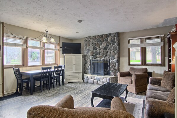 A spacious living room with a stone fireplace, dining area with chairs, and a television mounted on the wall.