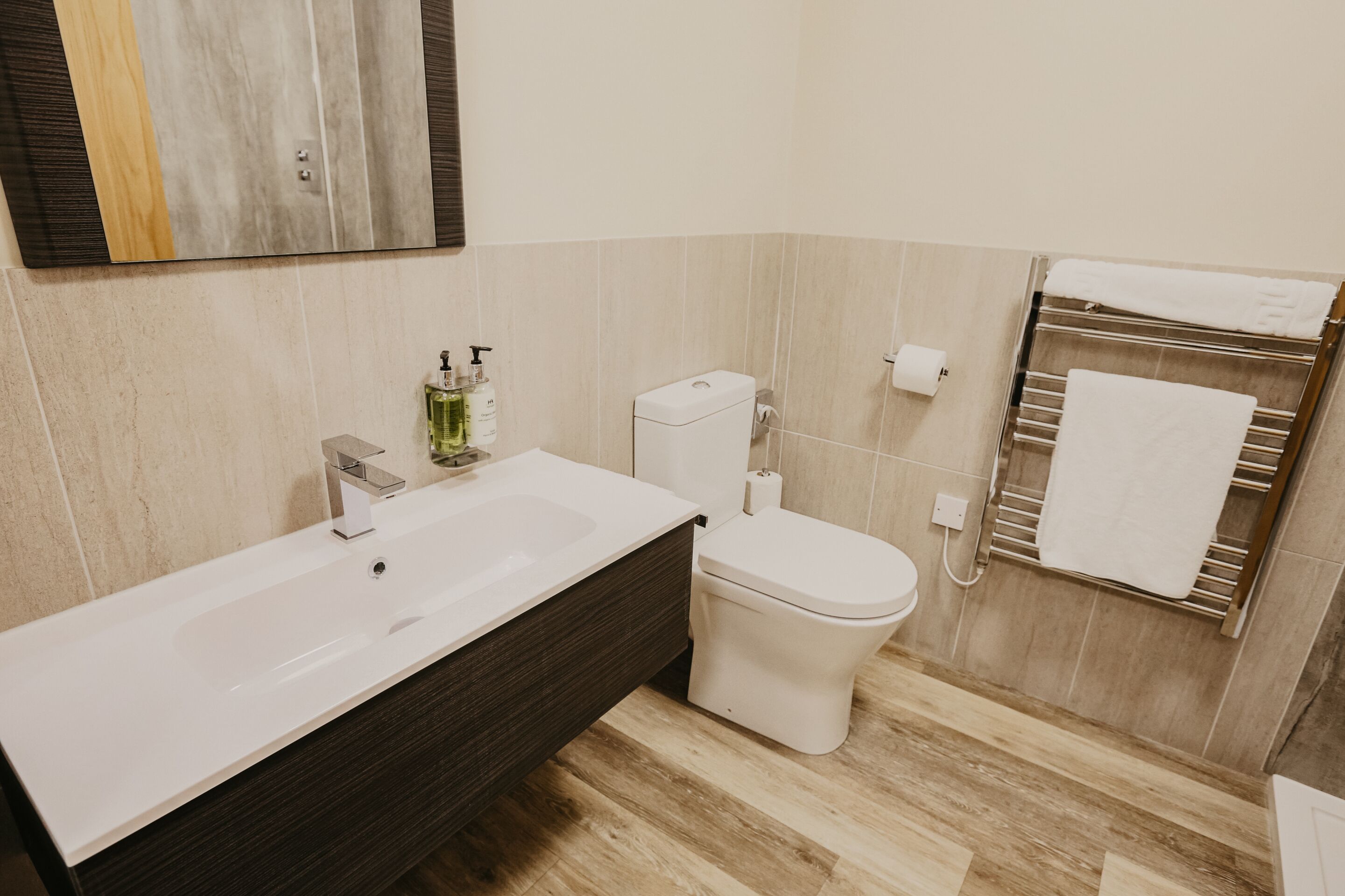 Et moderne badeværelse med en hvid vask, mørkt træ kommode, et hvidt toilet og en håndklædestang med hvide håndklæder.